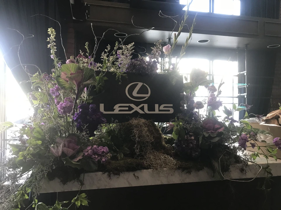 Moody floral installation framing a Lexus display, featuring deep purple and green tones, layered foliage, and organic textures arranged in a sculptural, asymmetrical composition.