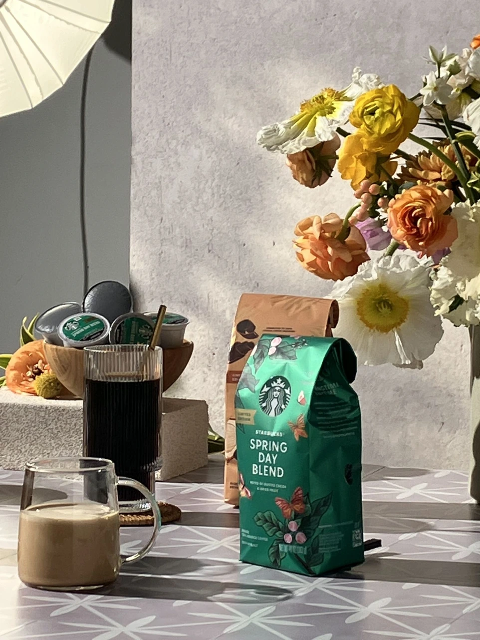 Styled coffee vignette with packaged Starbucks coffee, brewed drinks, and a spring floral arrangement featuring ranunculus and daffodils, arranged in soft natural light with a clean, modern backdrop.