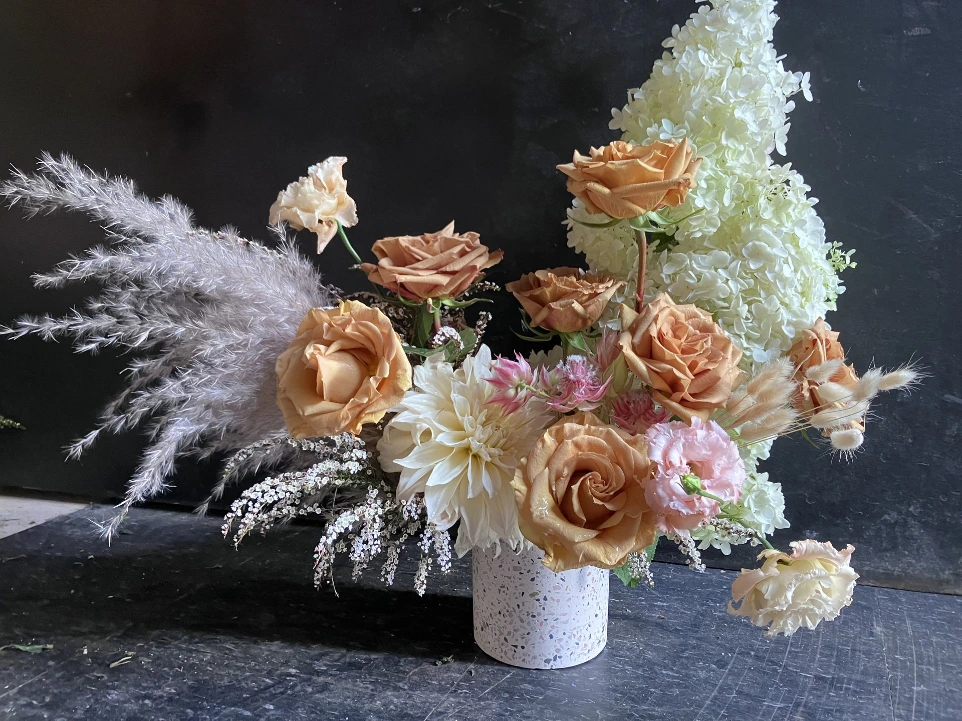 Textural floral arrangement in warm neutral tones featuring roses, hydrangea, lisianthus, and dried elements like pampas grass, arranged in an airy, sculptural composition.