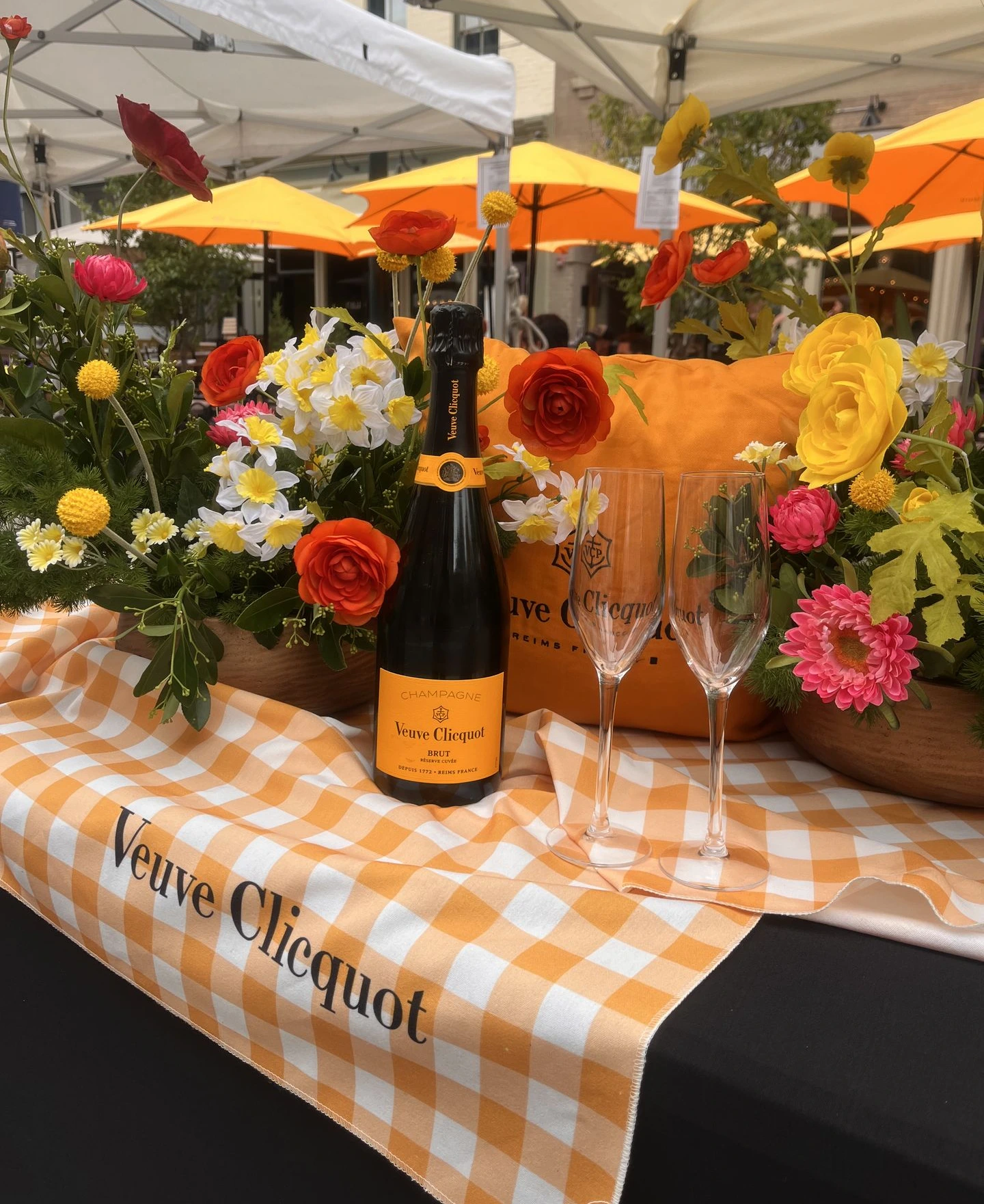 Outdoor event display with bright yellow, orange, and red floral arrangements surrounding a Veuve Clicquot champagne bottle and flutes, styled with branded linens and citrus-toned umbrellas for a bold, summery atmosphere.