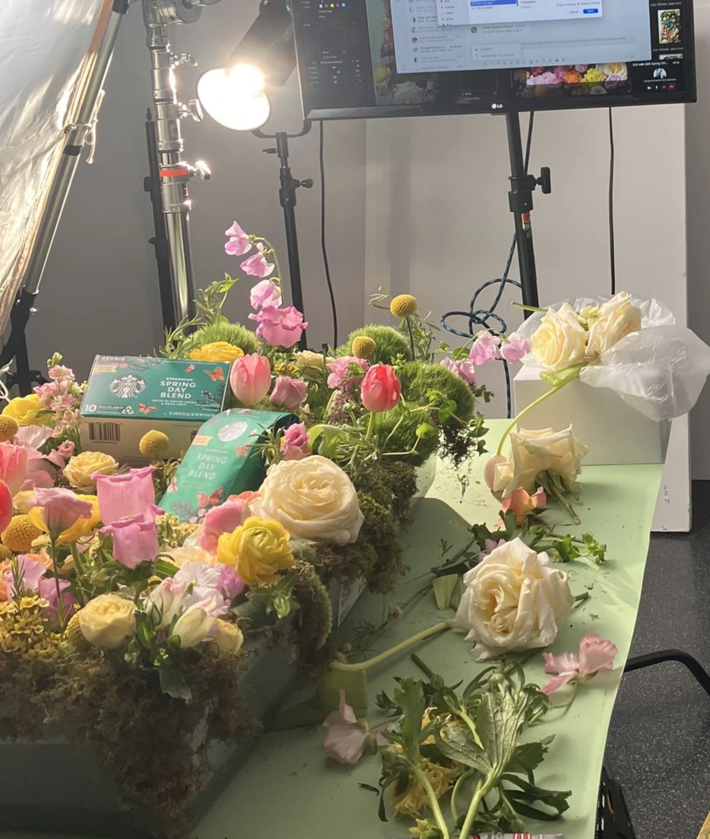 Behind-the-scenes floral styling setup with a worktable covered in fresh blooms, stems, and tools, illuminated by studio lighting as a designer reviews a live monitor during a product shoot.