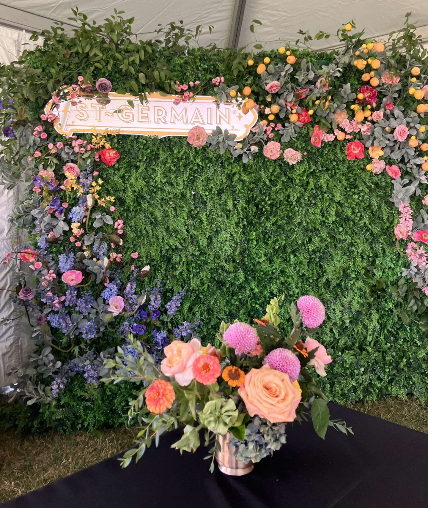 Lush event installation featuring a dense green hedge wall framed with colorful floral clusters in pink, coral, lavender, and yellow tones, accented with citrus elements and a St-Germain sign, with a bright, textural arrangement in the foreground.