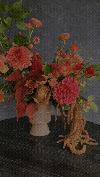 Artistic floral arrangement featuring coral dahlias, garden roses, textured foliage, and cascading amaranthus in a sculptural vase, designed by Juice Floral for luxury events and floral installations.